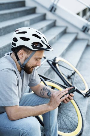 Photo for Young smiling bearded cyclist is sitting on the stairs with his bicycle nearby and looking at his phone. - Royalty Free Image