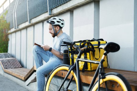Téléchargez les photos : Jeune messager de livraison express est assis sur un banc et la lecture d'un livre avec sac isolé et vélo à proximité. - en image libre de droit