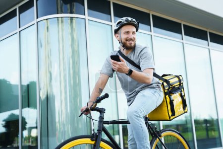 Photo for Young smiling express food delivery courier riding bicycle with insulated bag behind his back is holding his phone. - Royalty Free Image