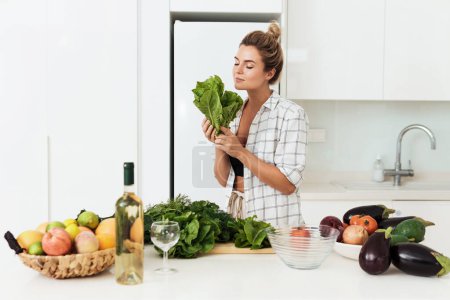 Téléchargez les photos : Jeune jolie femme avec renifler les feuilles de laitue pendant la cuisson dans la cuisine blanche - en image libre de droit