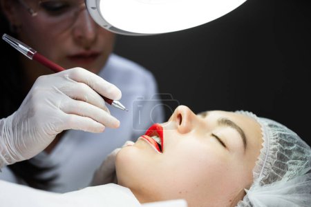 Téléchargez les photos : Maquilleuse professionnelle permanente et sa cliente pendant la procédure de rougissement des lèvres - en image libre de droit