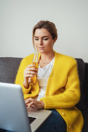 Foto de Mujer joven está sentada en el sofá bebiendo cerveza mientras ve películas o trabaja en su computadora portátil en casa - Imagen libre de derechos