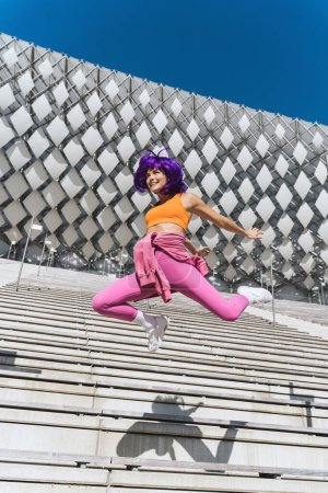 Téléchargez les photos : Jeune femme insouciante portant des vêtements de sport colorés sautant dans la rue pendant la journée d'été - en image libre de droit