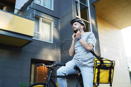 Téléchargez les photos : Jeune messager de livraison express avec sac isolé sur le dos met un casque de vélo tout en étant assis sur son vélo. - en image libre de droit