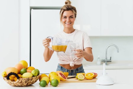 Photo for Young woman pouring freshly squeezed homemade orange juice into the glass in modern white kitchen - Royalty Free Image