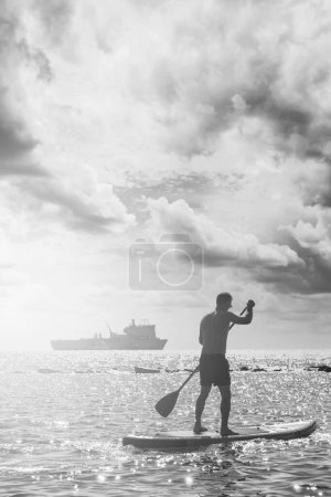 Téléchargez les photos : Jeune surfeur masculin chevauchant une planche à pagaie et ramant avec une pagaie dans un océan avec un navire en arrière-plan. - en image libre de droit
