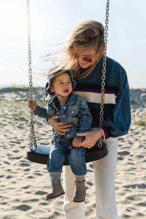 Téléchargez les photos : Jeune mère souriante tient son petit fils assis sur une balançoire à la plage de sable. - en image libre de droit