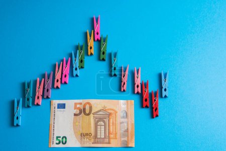 Téléchargez les photos : Gros plan d'un graphique financier en pinces à linge colorées en bois avec un billet de 50 euros en dessous sur fond bleu. - en image libre de droit