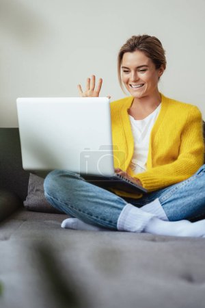 Foto de Joven mujer feliz saludando en la cámara portátil durante la conversación en línea por videollamada en casa - Imagen libre de derechos