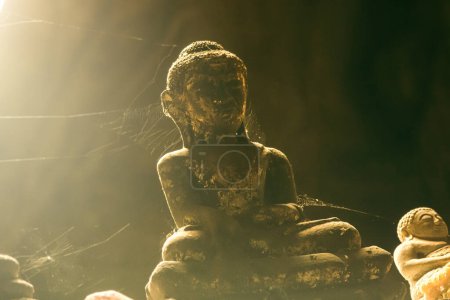 Téléchargez les photos : Gros plan d'une statue de pierre de Bouddha longtemps oubliée, recouverte de toile d'araignée, dans une grotte. - en image libre de droit