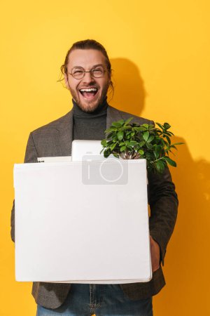 Téléchargez les photos : Jeune homme heureux tenant boîte avec des objets personnels après la promotion de l'emploi sur fond jaune - en image libre de droit
