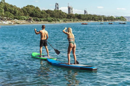 Photo for Couple of young male and female surfers are riding standup paddleboards together and rowing with paddles in the sea - Royalty Free Image