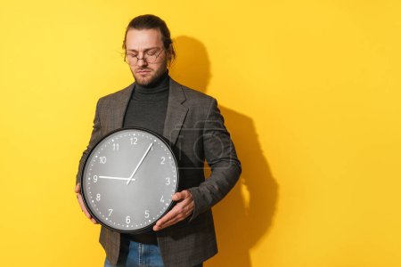 Photo for Sad bearded man wearing glasses holding big clock on yellow background - Royalty Free Image