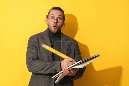 Téléchargez les photos : Homme barbu drôle portant des lunettes tenant grand crayon et cahier sur fond jaune - en image libre de droit