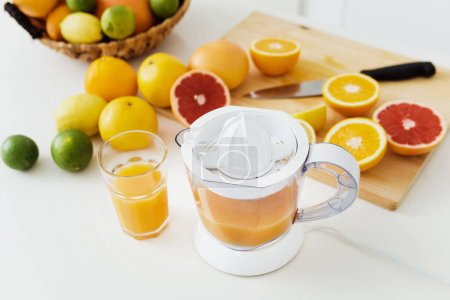 Foto de Vaso de zumo de naranja, exprimidor de cítricos y varios cítricos en mesa de cocina blanca - Imagen libre de derechos