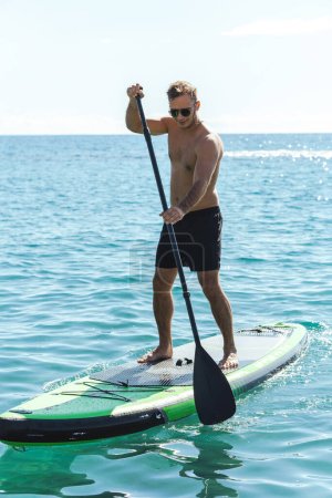 Young male surfer is riding a standup paddleboard and rowing with a paddle in an ocean.