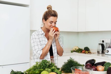 Téléchargez les photos : Jeune jolie femme avec renifler tomate mûre pendant la cuisson dans la cuisine blanche - en image libre de droit