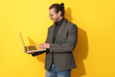 Photo for Handsome happy bearded man wearing eyeglasses is using laptop computer on yellow background - Royalty Free Image
