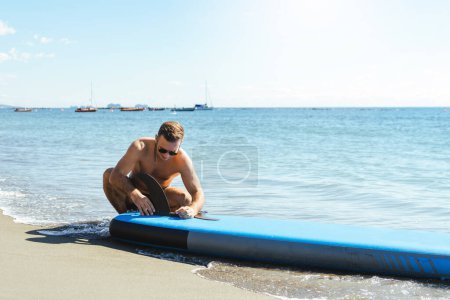 Photo for Young male surfer is setting up a standup paddleboard for a ride on a sandy beach near the ocean. - Royalty Free Image