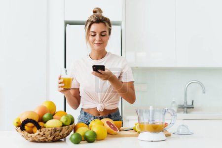 Photo for Young woman drinking freshly squeezed homemade orange juice and taking photos by her smartphone - Royalty Free Image