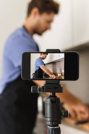 Photo for Young bearded chef blogger is cooking in the kitchen in front of a camera. - Royalty Free Image