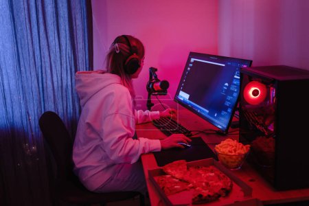 Photo for Young woman gamer or software developer sitting at the modern personal computer and eating junk food at night in room with neon lights - Royalty Free Image