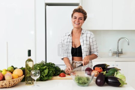 Foto de Mujer alegre joven cocina plato vegetariano saludable en casa en la cocina blanca - Imagen libre de derechos