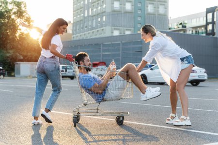 Photo for Three diverse friends having fun and riding shopping cart on parking lot near supermarket during summer evening - Royalty Free Image