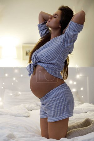 Téléchargez les photos : Jeune et sensuelle femme enceinte sur le lit dans son appartement confortable - en image libre de droit