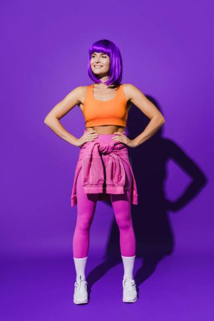 Téléchargez les photos : Portrait de femme joyeuse portant des vêtements de sport colorés assis sur fond violet - en image libre de droit