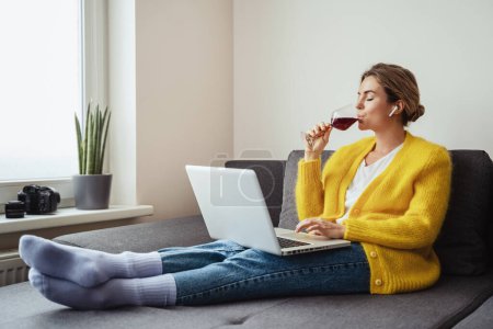 Junge Frau sitzt auf dem Sofa und trinkt Rotwein, während sie sich einen Film ansieht oder zu Hause an ihrem Laptop arbeitet