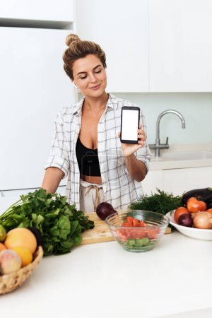 Foto de Mujer bonita joven está sosteniendo teléfono inteligente con pantalla en blanco para su diseño durante la cocina en la cocina moderna - Imagen libre de derechos