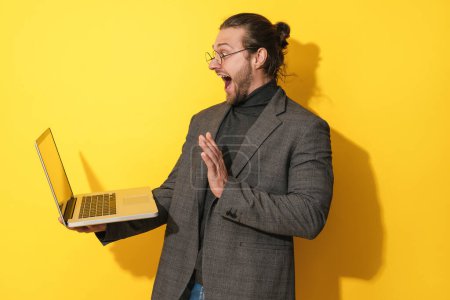 Photo for Surprised bearded man wearing eyeglasses waving into laptop screen during video call on yellow background - Royalty Free Image