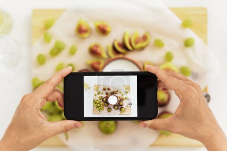 Foto de Mujer tomando fotos de su delicioso yogur griego con higos junto a su smartphone en cocina blanca - Imagen libre de derechos