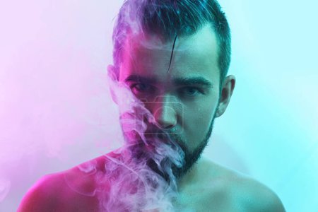 Portrait of handsome young man with wet skin in ultraviolet light smoking vape