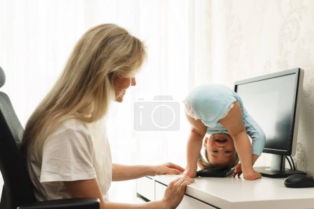 Photo for Little playful boy seeking his freelancer mother's attention is standing upside down on a desk at her workplace and distracting her from work on computer. - Royalty Free Image