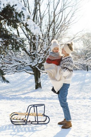 Foto de Joven madre y su lindo hijo pequeño con trineo retro en un parque nevado durante el soleado día de invierno - Imagen libre de derechos