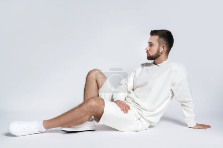Photo for Handsome man wearing white sweatshirt and shorts with wireless earbuds sitting against gray background - Royalty Free Image