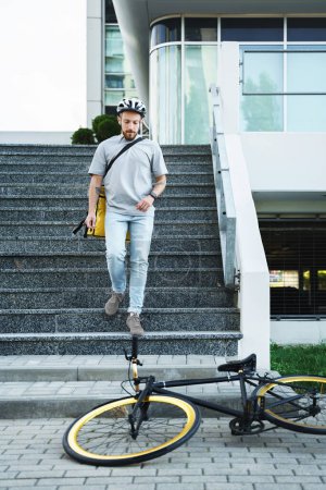 Photo for Young express food delivery courier with insulated bag behind his back is going down the stairs to his bicycle. - Royalty Free Image