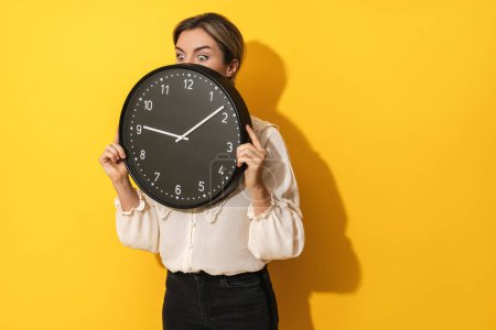 Foto de Joven empresaria asomándose detrás de un gran reloj sobre un fondo amarillo - Imagen libre de derechos