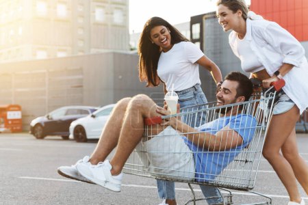 Photo for Three diverse friends having fun and riding shopping cart on parking lot near supermarket during summer evening - Royalty Free Image