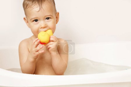 Téléchargez les photos : Adorable petit garçon met un canard en caoutchouc dans sa bouche tout en prenant un bain dans l'eau chaude. - en image libre de droit