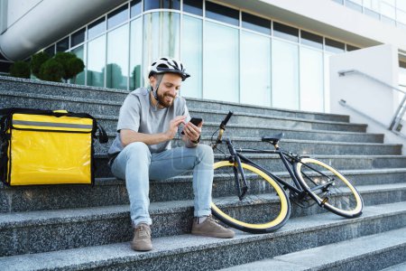 Photo for Young smiling express food delivery courier is sitting on the stairs with insulated bag and bicycle and looking at his phone. - Royalty Free Image