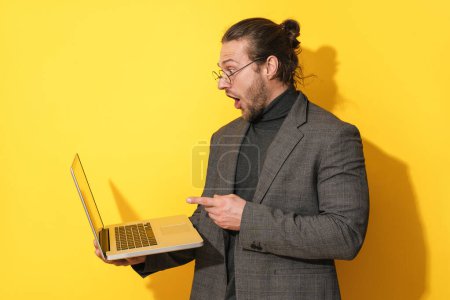 Foto de Sorprendido hombre barbudo con gafas apuntando a la pantalla del ordenador portátil en el fondo amarillo - Imagen libre de derechos