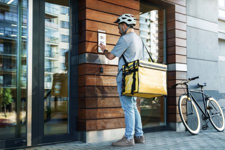 Téléchargez les photos : Jeune messager de livraison express avec sac isolé sur le dos utilise interphone avec vélo garé à proximité. - en image libre de droit