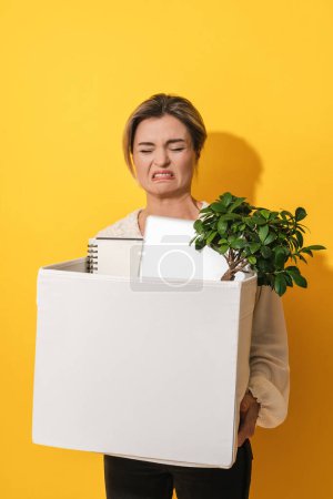 Photo for Young upset woman holding box with personal items after job resignation against yellow background - Royalty Free Image