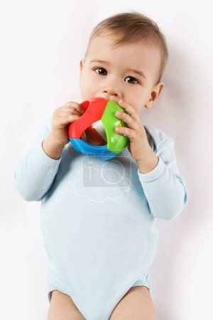 Foto de Adorable niño en un mameluco está acostado y jugando con un juguete de plástico, poniéndolo en su boca sobre un fondo blanco. - Imagen libre de derechos