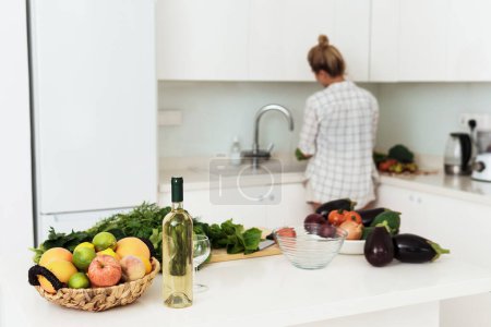 Foto de Botella de vino blanco, cesta de frutas y verduras diferentes en la mesa de cocina blanca - Imagen libre de derechos