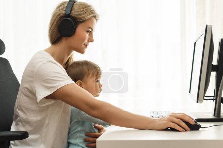 Foto de Joven madre freelancer está trabajando en la computadora en el lugar de trabajo de su oficina en casa con su pequeño hijo sentado en su regazo. - Imagen libre de derechos