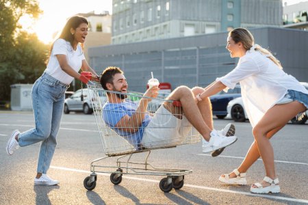 Foto de Tres amigos diversos que se divierten y montar carro de la compra en el estacionamiento cerca del supermercado durante la noche de verano - Imagen libre de derechos